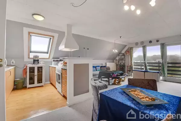 En åben plan stue med en stue til højre og et køkken til venstre. Et spisebord med blå dug står midt i rummet. Store vinduer giver udsigt til naturen udenfor.