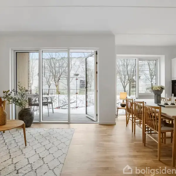 Stue med sofa og sofabord til venstre, spisebord og seks stole til højre. Store vinduer og glasdør åbner ud til en altan med udsigt til sneklædt gårdhave.