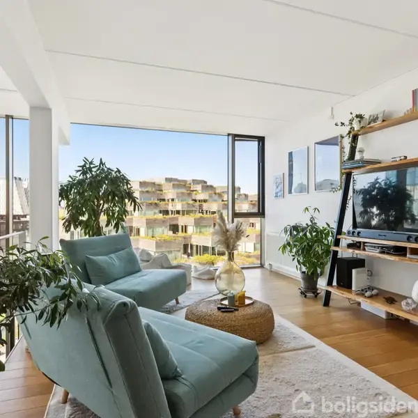 To lyseblå stole står i en moderne stue fyldt med grønne planter. En træreol holder et fjernsyn. Store vinduer giver udsigt over en by med sollys.
