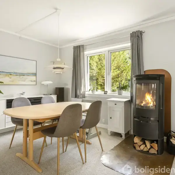 Et spisebord med fire stole står i et lyst rum ved siden af en brændeovn, der brænder. Vinduerne giver udsigt til grøn beplantning udenfor. Skabet og maleri fuldfører rummet.