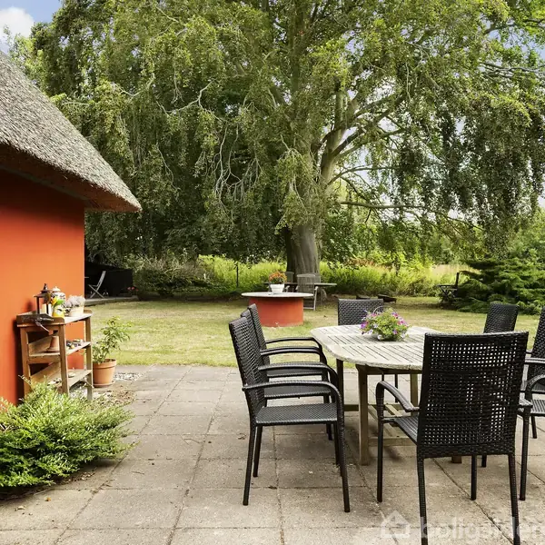 Spisebord med sorte stole står på en flisebelagt terrasse. Omgivelserne inkluderer et stråtækt, rødmalet hus og en frodig have med et stort træ i baggrunden.