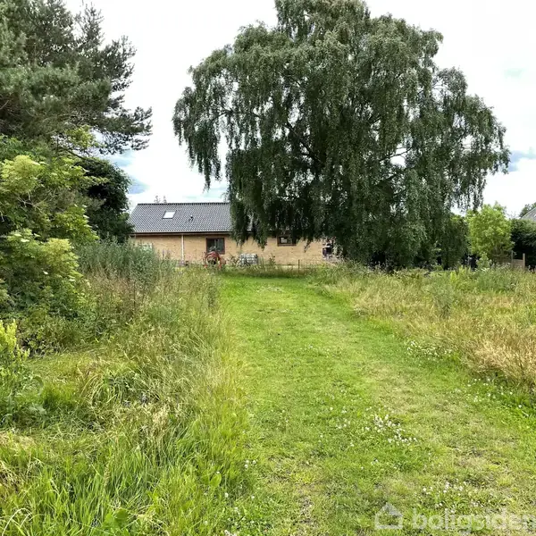 Et murstenshus står stationært i baggrunden, omgivet af en stor græsplæne med vildtvoksende grønne buske og et stort træ i centrum. Himmel med spredte skyer ses over.