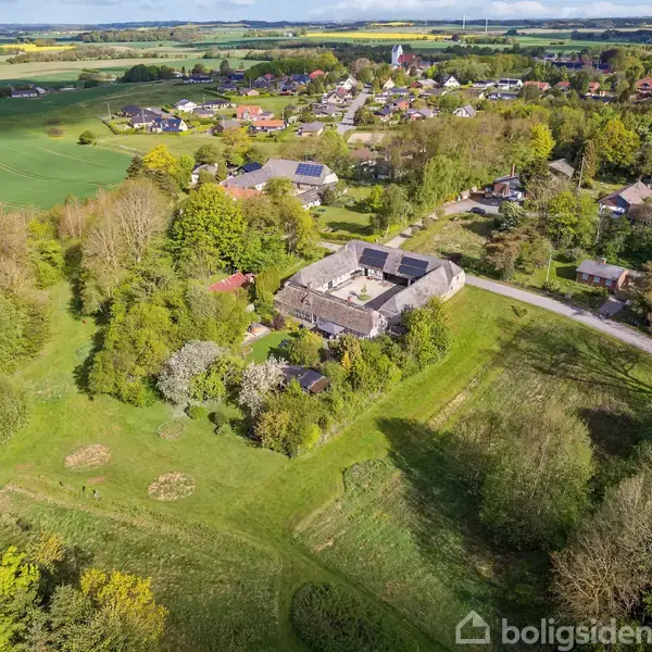 Landsby set ovenfra med spredte huse, marker i baggrunden, og en stor central bygning omgivet af træer. Solrigt vejr over et grønt, frodigt landskab.