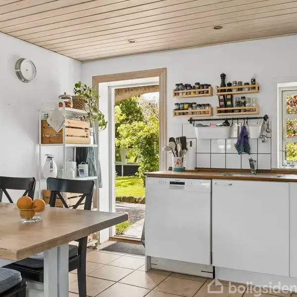 Køkken med hvidt interiør og træloft. Døre og vinduer åbner til en frodig have. Bord med sort stole, frugtskål og stearinlys. Hylder med krydderikler.
