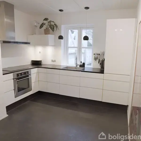 Et moderne køkken med hvide skabe og en indbygningsovn. Overfladen er sort, og der er en plante i hjørnet. Loftet har tre pendellamper. Vindue i baggrunden.