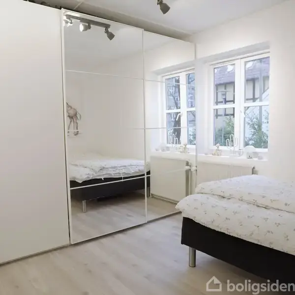 Et soveværelse med en seng dækket af et lyst sengetæppe. Skabet med spejllåger reflekterer værelset. Et vindue med udsigt til haven fylder rummet med naturligt lys.