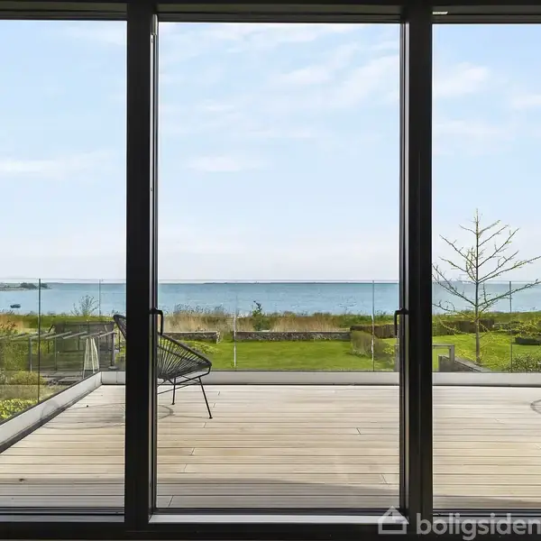 En stor glasskydedør åbner ud til en terrasse med udsigt over en frodig have og havet i baggrunden. To moderne stole står på terrassen.