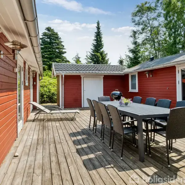 Rødt træhus med en træterrasse. Et bord med ti stole står midt på terrassen, omgivet af grønne træer og buske. Et par liggestole står ved siden af huset.