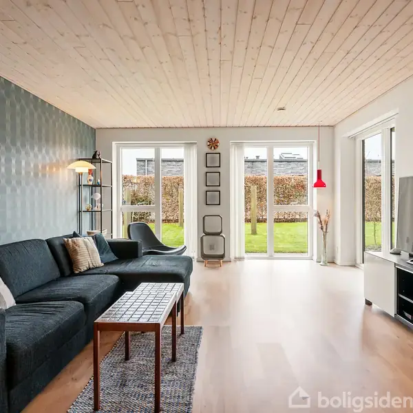 Stue med mørk sofa og sofabord foran et mønstret vægpanel, der åbner ud til en græsplæne gennem glasdøre. En reol og en lampe ved siden af sofaen.