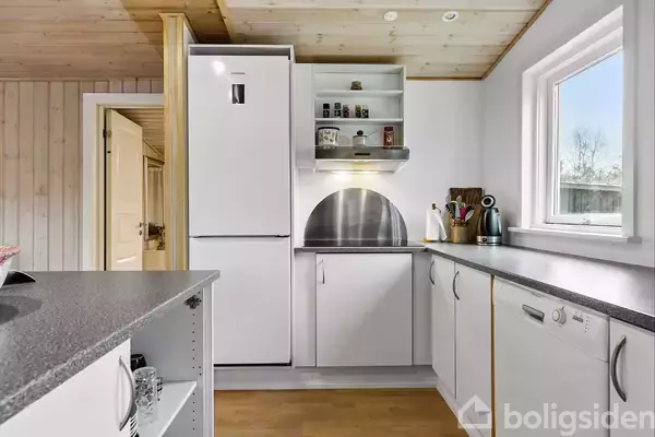 Et lyst køkken med hvidt køleskab, hylder og skabsplads. Bordpladerne er grå, og der er et vindue til højre, der giver naturligt lys. Træpaneler på væggene.