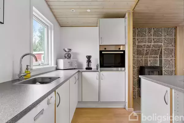 Et moderne køkken har hvide skabe med sorte bordplader. På bordet står en brødrister, blender og kaffemaskine. Væggene er lyse, og en komfur kan ses. En stor vindue giver lys.