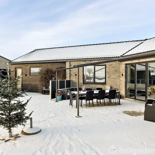 Et moderne hus med fladt tag står omgivet af sne i en gårdhave. Et bord med stole og et lille grantræ er synligt, omgivet af planter i krukker.