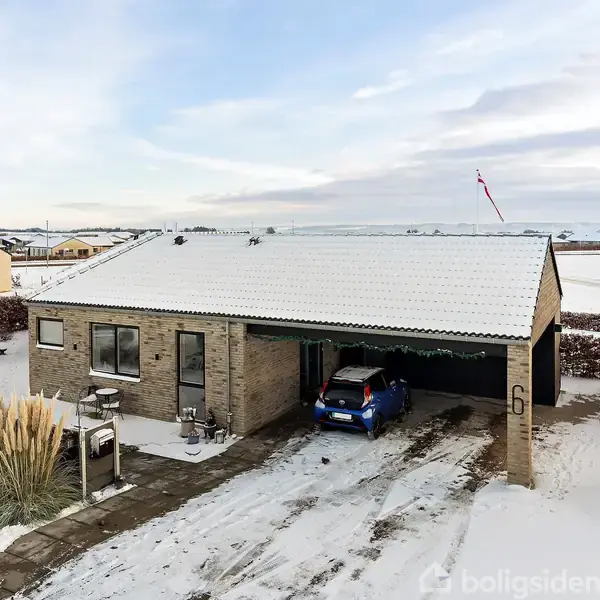 En enkeltstående, moderne murstenshus med garage, hvor en blå bil står parkeret. Sne dækker taget og omgivelserne. Numret "6" er synligt på væggen. Vinterlandskab med klare himmeltræk.