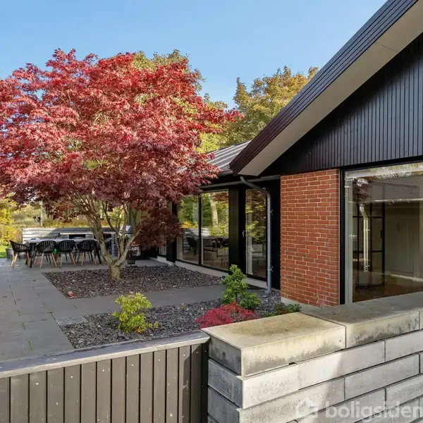 En moderne villa med røde mursten og sort træbeklædning står ved siden af en terrasse. Et træ med røde blade giver skygge over udendørs møbler i en have omkranset af mursten.