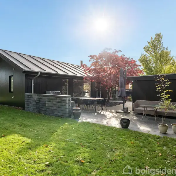 Sort hus med terrasse, der har havemøbler og parasol; placeret i en have med grønne træer og buske under en klar himmel med solskin.