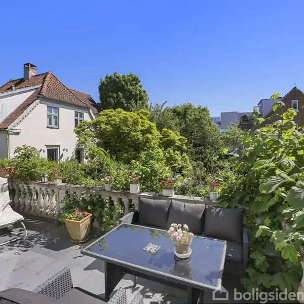 Et terrasseområde med sofabord og stole i et frodigt haveomgivelser. En charmerende hvid bygning med rødt tag ses i baggrunden under en klar blå himmel.