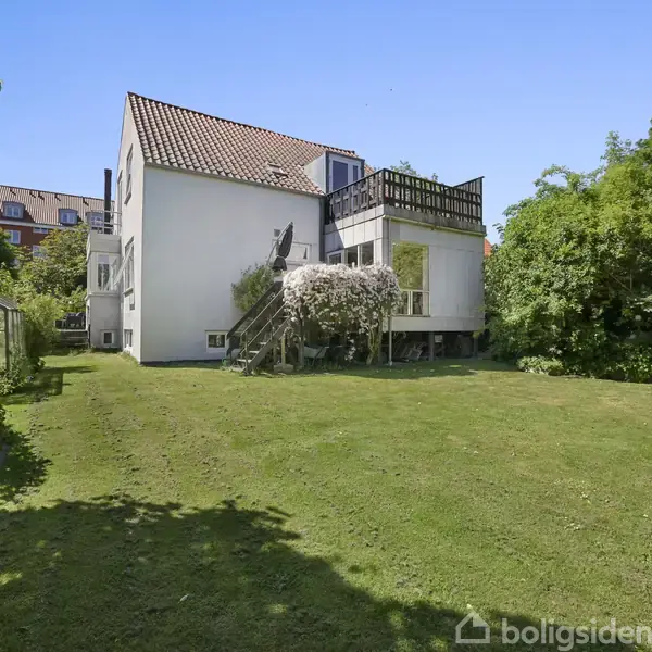 Huset står i en have med grøn græsplæne og omkranset af træer og buske. Der er en trappe op til en altan.