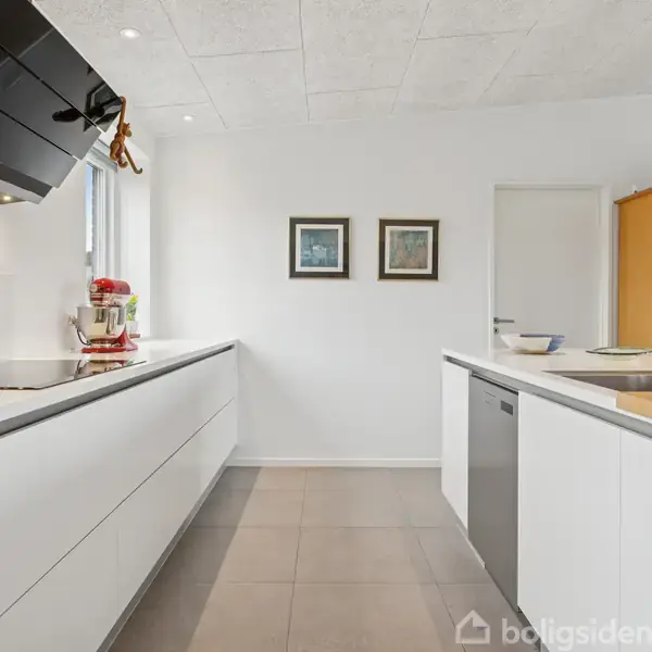 Moderne køkken med hvide skabe og stålarmatur. Køkkenmaskiner står på bordpladen. I baggrunden hænger to billeder på væggen. Rummet er lyst og minimalistisk.