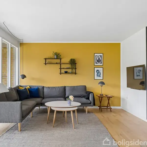Grå hjørnesofa står mod en gul væg, dekoreret med hylder og billeder. Rundt sofabord foran. Ved siden af er en væghængt pejs med brændestabel. Store vinduer til venstre.