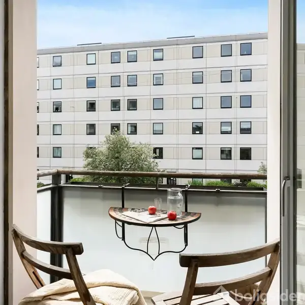 En altan med to træstole og et lille bord med glas og stearinlys. Set udefra er der en boligblok i baggrunden under en blå himmel.