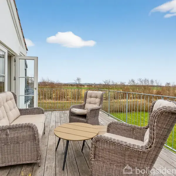 Tre kurvemøbler og et rundt bord står på en udendørs terrasse med udsigt over en græsplæne og natur i baggrunden. Den klare blå himmel understreger det lyse, rummelige udendørs område.