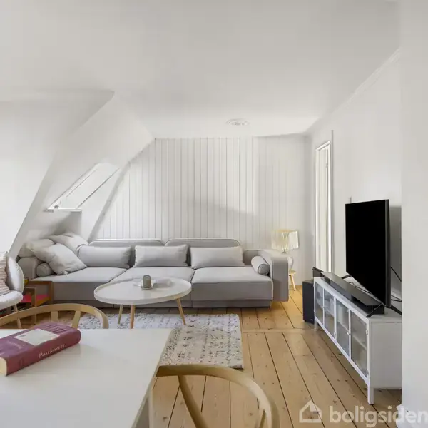 Grå sofa står mod hvid væg i lyst stue med trægulv, hvor der også er et tv, rund sofabord og reol. På spisebordet ligger en bog og en kop.