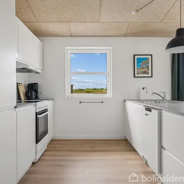 Et lyst køkken med hvide skabe og en indbygget ovn står i en rummelig, moderne indretning. Et vindue afslører en grøn udsigt, mens en sort lampe hænger fra loftet.