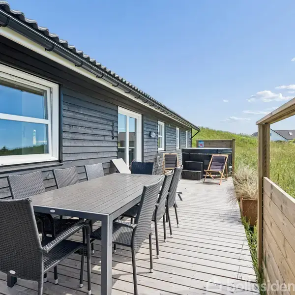 Sortmalet træhus veranda med spisebord og stole i flettet materiale. Foran er græs og buske, mens himlen er blå med få skyer.