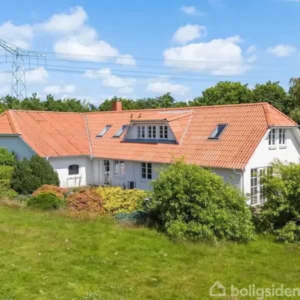 Et hvidt hus med rødt tegltag står omgivet af grønne buske og træer på en græsplæne under en blå himmel. Elektriske ledninger ses i baggrunden.