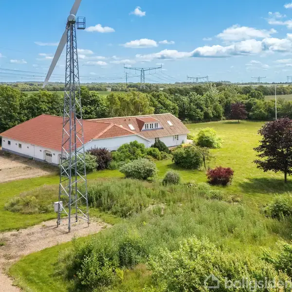 Hvid bygning med rødt tegltag står stille, omgivet af grønne marker og træer; en vindmølle står tæt ved siden i det åbne landbrugslandskab under en blå, skyet himmel.