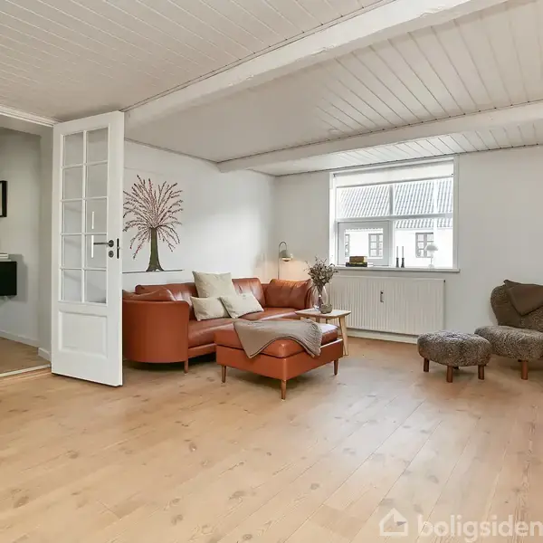 En stue med en brun lædersofa og en puff ved siden af. Rummet har trægulve, et stort vindue, og vægmonteret tv på højre side.