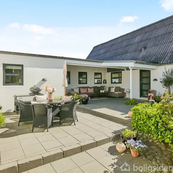 Terrasse med spisebord og stole står på fliser. I baggrunden er en hvid bygning med mørkt tag og vinduer. Omkring er der krukker med planter og en udendørs sofa under et halvtag.