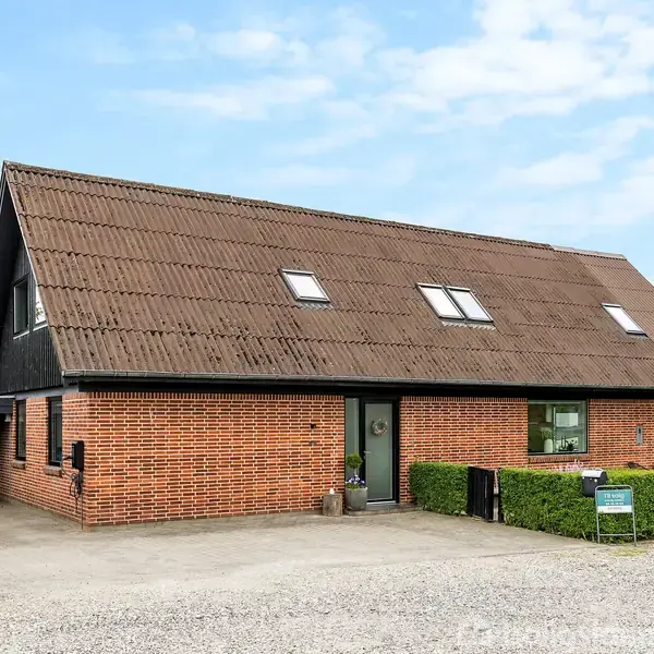 Et rødstenshus med skråtag og flere ovenlysvinduer står i en have med grusbelagt gårdsplads og grøn hæk. Huset har en sort træside og indgang ved flisebelagt sti.