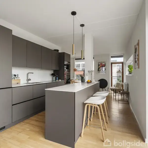 Moderne køkken med mørkegrå skabe, hvid bordplade og indbygget vask. Tre barstole står ved køkkenøen. I baggrunden ses spiseområde med lyst træbord og stole i en lys rumindretning.