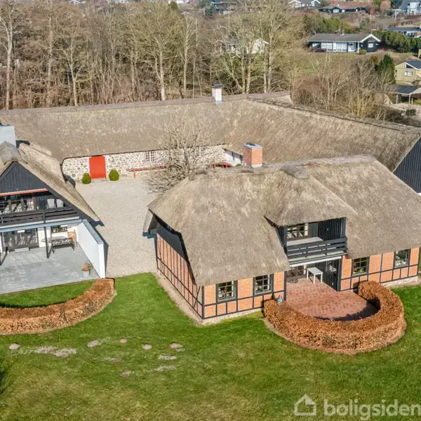 To stråtækte bygninger står på en grøn græsplæne, omgivet af lave hække og træer, med en indre gårdhave. Området er landligt og fredfyldt.