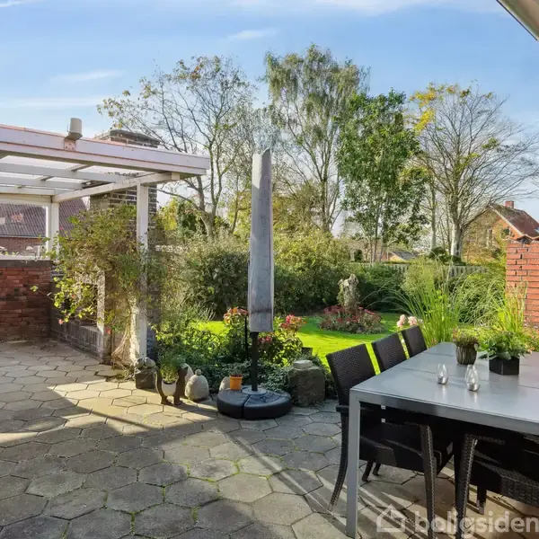 Terrassemøbler står placeret omkring et bord på en flisebelagt terrasse. En pergola giver skygge, mens en have med græs og beplantning omgiver området.