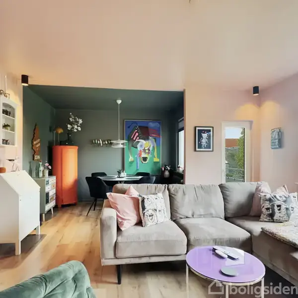 Grå sofa står centralt i moderne stue med trægulv, ved siden af et lilla sofabord. I baggrunden ses en spisestue med farverig kunst på væggen.
