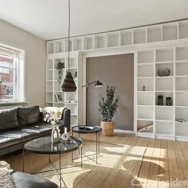 Sort læderhjørnesofa står foran et stort vindue i en lys, moderne stue. Rundt sofabord og væghylder omkranser et indbygget skrivebord med dekorative genstande, på trægulv.