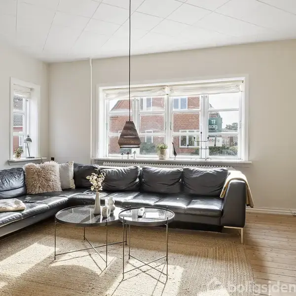 Sort læderhjørnesofa står i en lys stue med store vinduer. To runde glasborde foran sofaen, planter i hjørnet, trælaminatgulv og hvidt loft.