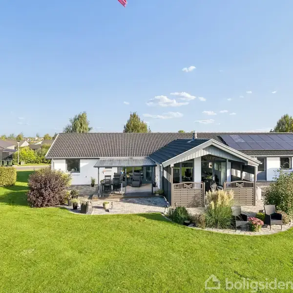 Hus med sort tag, solpaneler og en stor terrasse i en velplejet have. Hækken omkranser området, og himlen er klar med få skyer.