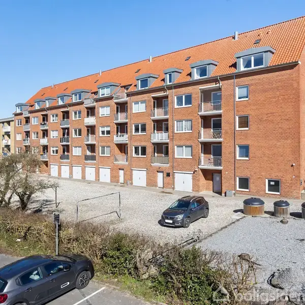En stor, rød murstensejendom står i en parkeringsplads med biler parkeret foran. Ejendommen har flere etager med små altaner og et skråt, rødt tegltag.