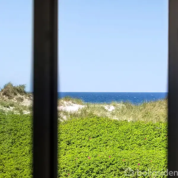 Havudsigt gennem et vindue; bølgerne skimtes i det fjerne over grønne klitlandskaber. Klar blå himmel danner baggrunden.