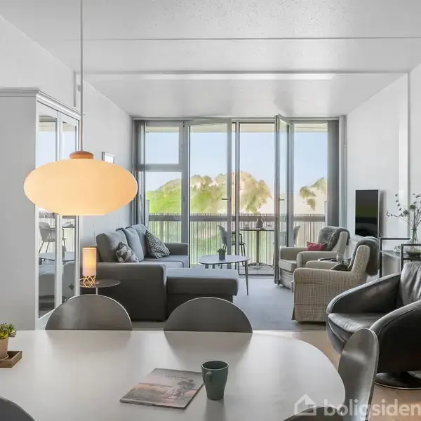 Stuen har moderne møbler, herunder sorte læderstole og grå sofa, vendt mod store vinduer, der giver udsigt til en balkon og grønne omgivelser. En rund lampe hænger over bordet.