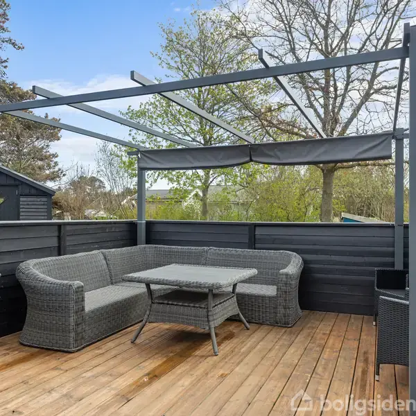 En grå sofa og bord står under en metalramme på en træterrasse. Træer og grønne omgivelser ses i baggrunden.