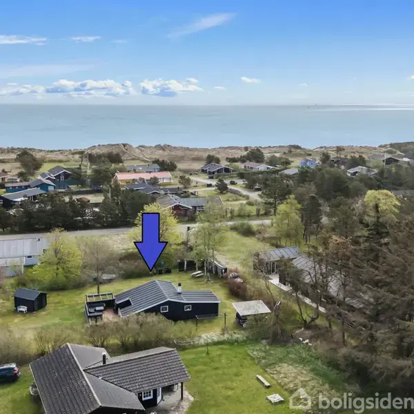 Et sommerhus er markeret med en blå pil og ligger i et grønt område nær kysten. Flere huse omgiver det, med havudsigt i baggrunden.
