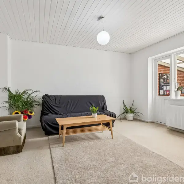 Sofa dækket med sort betræk står ved siden af et træbord i en lys stue. En beige lænestol og planter dekorerer rummet. Sollys strømmer ind gennem store vinduer.