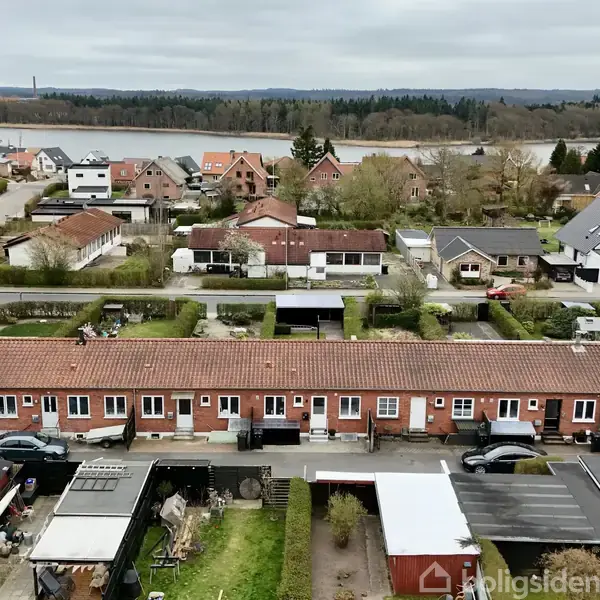Rækkehus omgivet af villakvarter med haver og veje, set fra oven. Biler er parkeret foran husene. I baggrunden er der en sø og skovområde.