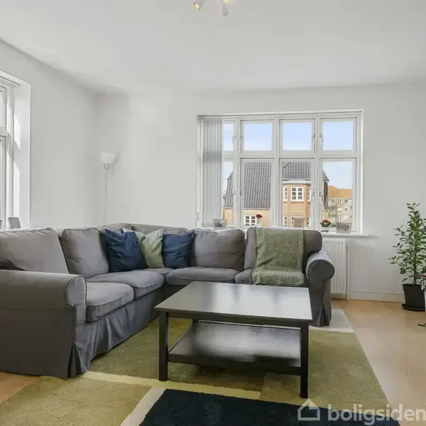 Grå sofa står i hjørnet af en lys stue, med et sort sofabord foran og grønne planter i nærheden; store vinduer slipper lys ind, mens gardiner hænger til siden.