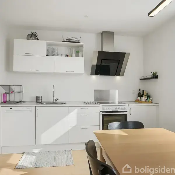 Et lyst køkken har hvide skabe, induktionskomfur og emhætte. Ved siden af står et spisebord med sorte stole. Et vindue lader lys ind på rummets hvide vægge.