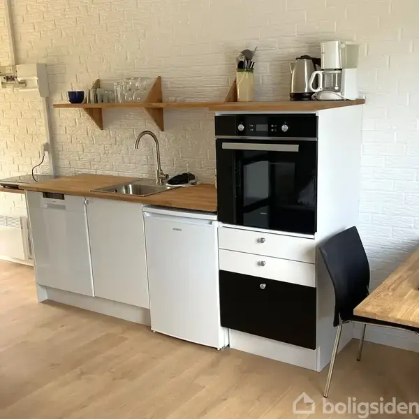 Et lille køkken med en ovn og kogeplade integreret i et skab. En vask ved siden af er monteret i en træbordplade. Der er åbne hylder med kopper og glas over bordpladen. En spiseplads med et bord og en stol står til højre.